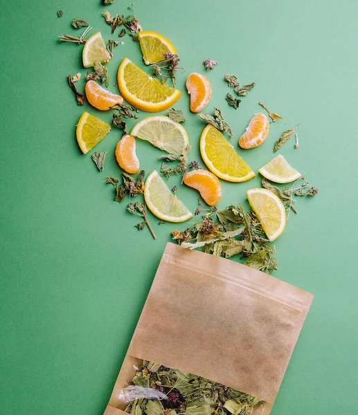 Leaf tea spilled out of a paper packet with orange and tangerine slices on green background