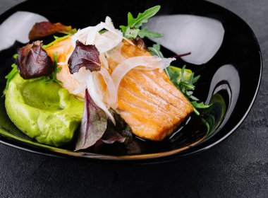 Close-up of grilled salmon fillet with green arugula sprouts with mashed avocado on a plate.