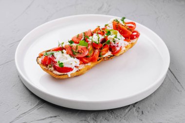 Lezzetli bruschetta, olgun domatesler, peynir ve otlar beyaz tabakta, gri arka planda.