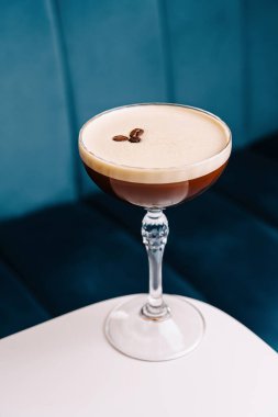 Kahve çekirdeği garnitürlü espresso martini, beyaz bir masada mavi bir arkaplanda servis edilir.