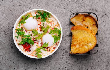 Yoğurt üzerinde haşlanmış yumurta, otlarla süslenmiş, çıtır tostla servis edilen bir kasenin üst görüntüsü.