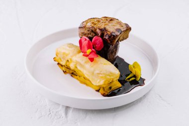 Nefis pişmiş biftek ve yenilebilir çiçeklerle süslenmiş katmanlı patates, beyaz bir tabakta servis edilir.