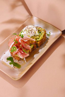 Pürüzsüz jambonlu nefis avokado tostu, haşlanmış yumurta ve şık bir tabakta taze otlar.