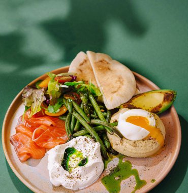 Sağlıklı kahvaltı tabağı, haşlanmış yumurta, tütsülenmiş somon, avokado ve yeşil arka planda taze yeşiller.
