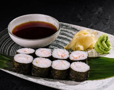 Wasabi, zencefil ve bambu yaprağına batırılmış soslu suşi rulolarının zarif sunumu ve desenli siyah arka plan.