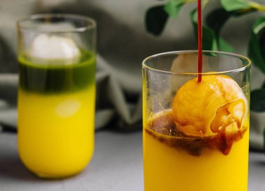 Canlı turuncu şerbet bir bardak meyve suyuna dökülüyor, şık gri bir arka planda.