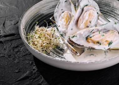 Kremalı sosa batırılmış, lahanayla süslenmiş, koyu bir arka planda şık bir tabakta sunulan leziz midyeler.