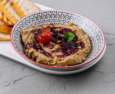 Üstü otlarla kaplı kremalı humus tabağı ve yanında kızarmış domates, yanında da kızarmış ekmek.