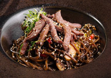 Lezzetli, sebzeli ve erişteli kızarmış biftek lahanayla süslenmiş, siyah bir tabakta servis edilir.