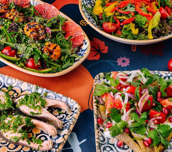 Arkaplanda taze malzemelerle desenli bir masa örtüsü üzerinde dekoratif kâselerde servis edilen canlı Akdeniz mutfağı.