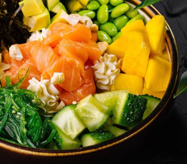 Siyah arka planda somon, mango, avokado ve edamame içeren canlı bir poke kasesi.
