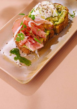 Pürüzsüz jambonlu nefis avokado tostu, haşlanmış yumurta ve şık bir tabakta taze otlar.