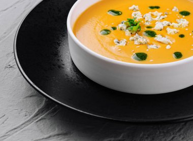 Bitki ve tohumlarla süslenmiş kadife kabak çorbası, siyah bir tabakta şık bir kasede servis edilir.