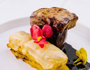 Nefis pişmiş biftek ve yenilebilir çiçeklerle süslenmiş katmanlı patates, beyaz bir tabakta servis edilir.