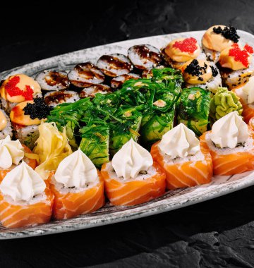 Çeşitli suşi dürümleri ve Nigiri çeşitleri modern bir tabakta koyu bir arkaplanda sunuluyor.