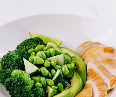 Beyaz bir tabakta taze brokoli, bezelye ve avokado ile ızgara tavuk dilimleri ile besleyici bir yemek.