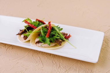 Pürüzsüz beyaz bir tabakta roka ve kurutulmuş domatesle süslenmiş zarif etli tacolar.