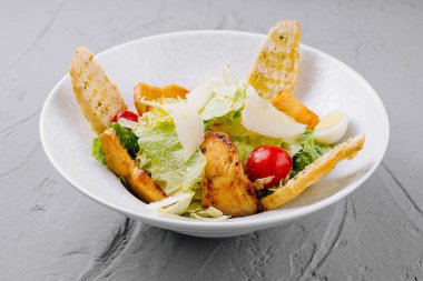 Bir kase sezar salatası ızgara tavuk, kızarmış ekmek ve gri arka planda vişneli domates.