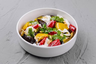 Colorful salad with grilled vegetables, herbs, and cheese in a white bowl on a grey background