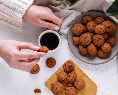 Close-up of hands holding a cookie with a cup of coffee and a plate of cookies on a white table