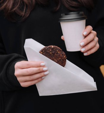 Close-up of a woman in a black jacket holding a tasty cookie and a takeaway coffee cup