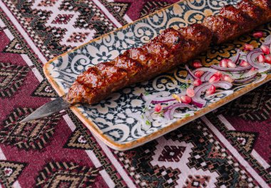 Dilimlenmiş soğanla dekoratif bir tabakta servis edilen sulu kebap geleneksel desenli bir kumaşta sunuluyor.