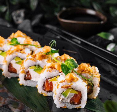 Canlı malzemelerle dolu sanatsal bir suşi sunumu ve taşların ve yaprakların arasında parlak, koyu bir tabakta garnitür.