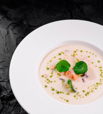 Taze otlarla dolu lüks deniz ürünleri çorbası siyah desenli beyaz bir tabakta sanatsal bir şekilde sunuluyor.