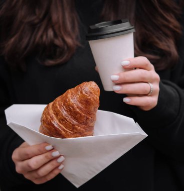 Close-up of a woman's hands with a coffee cup and a fresh croissant wrapped in paper