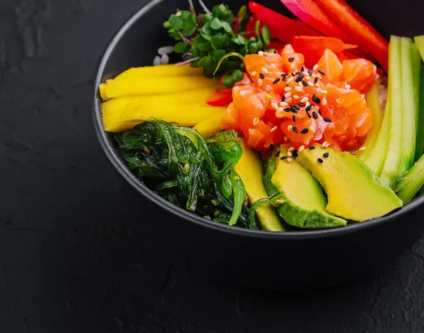 stock image Colorful poke bowl with salmon, mango, avocado, and vegetables on a dark slate surface