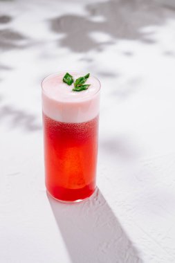 Kırmızı kokteyli köpük ve fesleğen yapraklarıyla tazeleyerek beyaz arka planda gölgelerle dururlar.