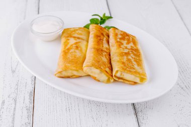 Tahta bir masada beyaz bir tabakta içi doldurulmuş ince krepler, bir tutam nane ile süslenmiş.