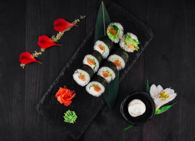 Somon ve avokado ile taze suşi ruloları çiçeklerle süslenmiş siyah taş servis tabağında servis edilir.