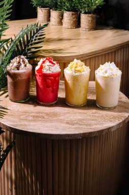 Çikolata, çilek, mango ve vanilya aromalı dört milkshake, krem şanti ve taş bir masanın üzerinde duruyor.