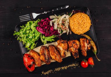 Siyah bir tabakta servis edilen ızgara tavuk kanatları marul, lahana, soğan, bulgur, domates, biber ve siyah ahşap arka planda otlarla servis edilir.
