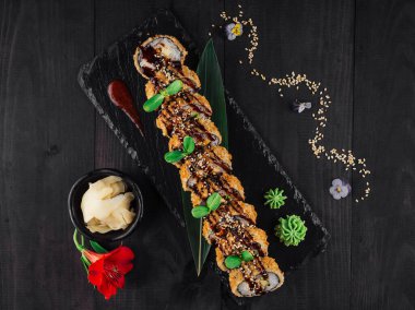 Çiçeklerle süslenmiş Tempura maki suşi ruloları ve susam tohumları siyah bir taş tabakta sunuluyor, turşulu zencefil ve wasabi ile.
