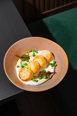 Siyah bir masada bej bir tabakta duran yeşil bezelyeli tavuk pirzolası ve beyaz sos.