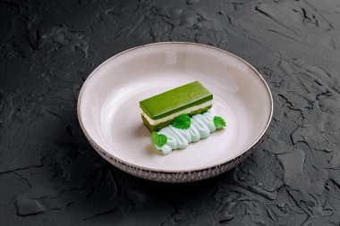 Beyaz çikolata ve nane kremalı yeşil çay dilimi, siyah desenli arka planda beyaz bir tabakta servis edilir.