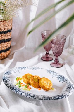 Beyaz tabakta servis edilen yuvarlak patates krepleri çiçek desenli, beyaz sos ve beyaz masa örtüsü üzerine yeşiller.