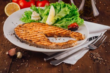 Izgara somon bifteği, ahşap bir masada marul, limon ve baharatla birlikte beyaz bir tabakta servis edilir.