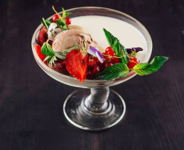 Lezzetli dondurma tatlısı, taze meyve ve nane kokulu yapraklarla süslenmiş zarif cam bir vazoda sunuluyor.