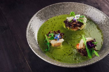 Karidesli yeşil krema çorbası, roka, çiçekler ve zeytinyağı. Koyu kırsal bir ahşap masada bir restoranda servis ediliyor.