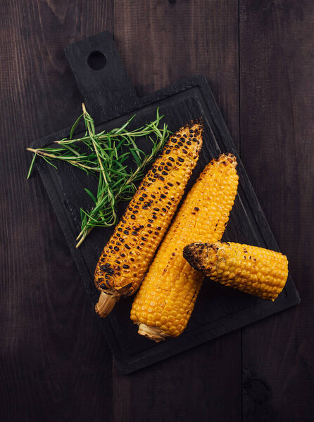 Жареная кукуруза на початке лежит на черной доске и темном деревянном столе, украшенном свежим розмарином