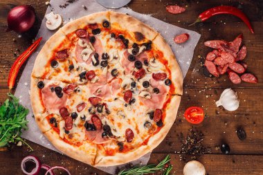 Eritilmiş mozzarella, jambon, mantar ve siyah zeytinli ağız sulandıran pizza, taze malzemelerle çevrili, kırsal ahşap bir masanın üzerinde yatıyor.