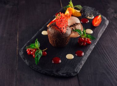 Çikolatalı kek ve çikolatalı dondurma siyah bir tahta üzerinde sunulur. Meyve ve sosla süslenir.