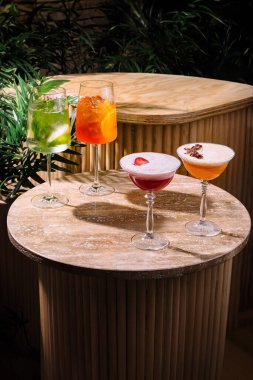 Çeşitli süslerle dört kokteyl bir masanın üzerinde duruyor. Yakındaki bitkilerden süzülen benekli güneş ışığıyla aydınlatılıyor.