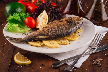 Beyaz tabakta ızgara balık, patates, domates, limon, marul ve baharatla ahşap masada servis edilir.