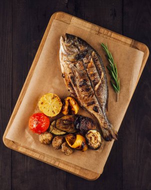 Koyu ahşap masada limonlu, biberiyeli ve sebzeli ızgara dorado balığı.