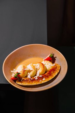 Çilek, badem ve sosla süslenmiş karamelize edilmiş muz üzerinde süzme peynir krepleri servis edilir.