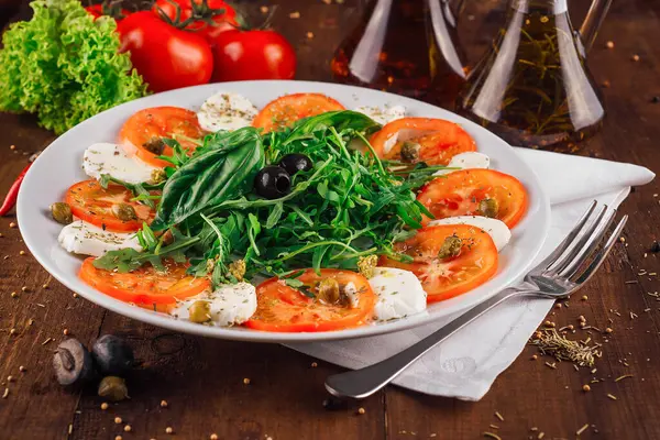 Domatesli taze Caprese salatası, mozzarella peyniri ve zeytin yağı ve baharatla tatlandırılmış fesleğen, ahşap bir masada beyaz bir tabakta servis ediliyor.